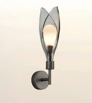 A modern wall sconce light fixture with a dark metal wall mount and a translucent smoky glass shade shaped like stylized leaves, shown illuminated against a soft beige background.