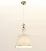 A modern pendant light with a textured off-white fabric drum shade, a small pale pink decorative bead, and brass-finished metal hardware including a ceiling canopy and top cap, shown against a plain off-white background.