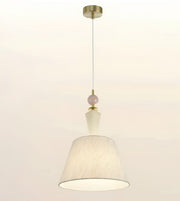 A modern pendant light with a textured off-white fabric drum shade, a small pale pink decorative bead, and brass-finished metal hardware including a ceiling canopy and top cap, shown against a plain off-white background.