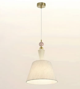 A modern pendant light with a textured off-white fabric drum shade, a small pale pink decorative bead, and brass-finished metal hardware including a ceiling canopy and top cap, shown against a plain off-white background.