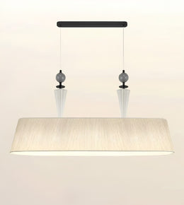 A modern rectangular pendant light fixture with a wide, tapered beige fabric shade, suspended from a black horizontal bar with two clear glass cone drops and dark gray spherical accents.