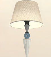 A modern table lamp with a pleated beige fabric drum shade, a light blue glass sphere, and sculptural white and gray geometric bases on a warm off-white background.