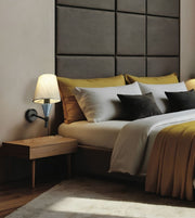 A modern bedroom scene featuring a bed with white and mustard yellow bedding, dark gray pillows, a dark gray padded headboard, a wooden bedside table, and a lit wall lamp.