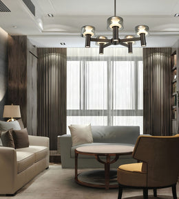 A modern living room interior featuring a multi-light chandelier with cylindrical glass shades, a light gray sofa, a cream sofa, a round wooden coffee table, and a brown armchair with a yellow cushion, set against dark wood paneling and sheer curtains.