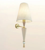 A modern wall-mounted sconce with a tapered off-white fabric shade, a white ceramic cone body, and brass-finished metal hardware, shown against a plain light beige background.
