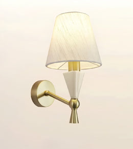 A modern wall-mounted sconce with a tapered off-white fabric shade, a white geometric cone detail, and a brushed gold metal arm and wall plate, shown against a plain light background.