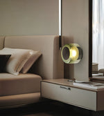 A modern wall-mounted cylindrical lamp with a glowing warm yellow light, set in a sleek contemporary bedroom next to a beige bed and floating nightstand.