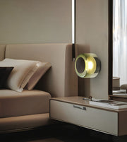 A modern wall-mounted cylindrical lamp with a glowing warm yellow light, set in a sleek contemporary bedroom next to a beige bed and floating nightstand.
