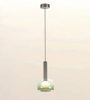 A modern single pendant light with a brushed metal canopy and rod, featuring a frosted glass shade that glows with a warm yellow light, set against a soft beige gradient background.