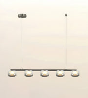 A modern 5-light linear pendant chandelier with clear glass cylindrical shades and a dark metal frame, suspended from a ceiling canopy against a light beige background.
