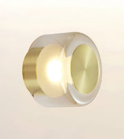 A modern cylindrical wall sconce with a brushed gold metal body, featuring a glowing warm white light in the center and a polished gold circular face on the right, set against a soft beige gradient background.