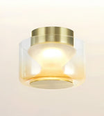 A modern flush-mount ceiling light fixture with a brushed gold metal top canopy and a clear glass shade that fades to frosted white, glowing with a warm yellow light against a clean off-white background.