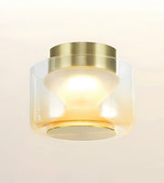 A modern flush-mount ceiling light fixture with a brushed gold metal top canopy and a clear glass shade that fades to frosted white, glowing with a warm yellow light against a clean off-white background.