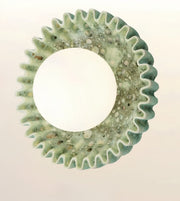 A circular decorative wall mirror with a textured, scalloped outer frame made of light green resin and embedded small pebbles, set against a plain off-white background.