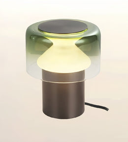 A modern table lamp with a translucent smoky green glass shade, a glowing warm white frosted center, and a matte dark bronze cylindrical base on a light beige background.