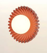 A circular wall sconce with a fluted, sunburst-like outer rim in terracotta orange-brown, featuring a large white illuminated center on a light beige background.