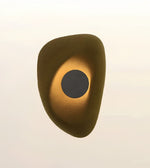An abstract organic-shaped wall sconce or decorative light fixture with a textured metallic gold finish and a dark circular center, shown against a plain off-white background.