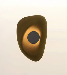 An abstract organic-shaped wall sconce or decorative light fixture with a textured metallic gold finish and a dark circular center, shown against a plain off-white background.