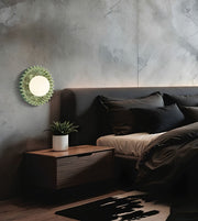 A modern bedroom scene featuring a decorative green sunburst wall mirror mounted on a textured gray concrete wall above a dark wood nightstand and bed.