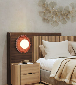 A modern bedroom scene featuring a wooden headboard with a built-in circular orange flower-shaped night light, a matching light wood nightstand, and a bed dressed in white and beige linens.