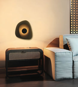 A modern wall-mounted sculptural lamp with a dark organic shape and glowing circular center, shown in a styled living room next to a light blue sofa and a dark side table.