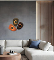 Modern living room setting featuring a three-piece abstract wall art set with glowing circular centers, mounted on a textured gray wall above a light beige sectional sofa.