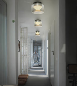 Three modern flush-mount drum ceiling lights with brushed silver metal tops and white shades, illuminated in a hallway with white walls and a hanging chair in the distance.