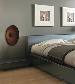 A modern low-profile platform bed with a dark gray headboard and matching side table, styled with beige and gray bedding. The room features a gray wall with two white square frames containing textured beige leaf designs, and a copper-colored circular wall decor piece on the left.