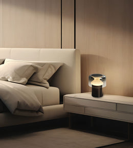A modern bedside table lamp with a glowing warm bulb inside a clear dome and dark base, placed on a light beige nightstand next to a matching bed in a minimalist bedroom.