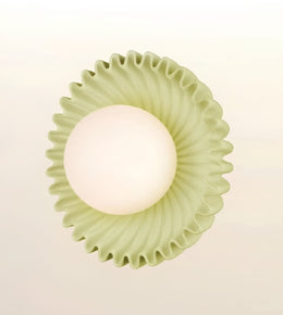 A modern wall-mounted light fixture with a textured, fluted circular shade in a pale sage green and a glowing frosted white globe bulb, shown against a soft off-white background.