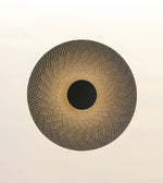 A circular woven basket or mat with a textured geometric pattern, featuring a dark brown center and a light brown outer rim, set against a plain off-white background.