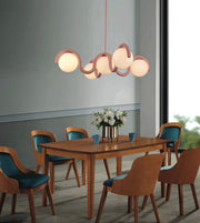 A modern dining room scene featuring a wavy, multi-light pendant chandelier with glowing frosted glass globes and a warm wood frame, hanging above a wooden dining table and six matching chairs with teal cushions.