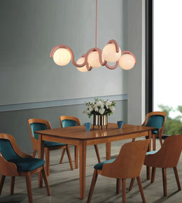 A modern dining room scene featuring a wavy, multi-light pendant chandelier with glowing frosted glass globes and a warm wood frame, hanging above a wooden dining table and six matching chairs with teal cushions.