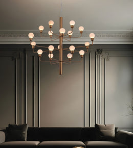 A modern multi-light chandelier with a brushed brass/gold metal frame and frosted white globe bulbs, hanging in a styled living room with a dark gray wall, decorative molding, and a dark sofa below.