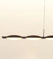 A modern linear pendant light with three illuminated oval frosted shades and a dark metal frame, suspended by a thin cord against a plain off-white background.