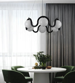 A modern black wavy pendant light with five white spherical glass shades hangs over a round white dining table with green chairs in a contemporary room with sheer white curtains and dark gray drapes.