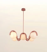 A modern wavy pendant light fixture with a dusty rose metal frame and five frosted glass globes, suspended from a matching ceiling canopy against a soft off-white background.