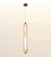 A modern vertical pendant light with a brushed brass canopy and cord, featuring two stacked cylindrical glass shades with a warm glowing light inside, set against a plain off-white background.