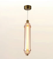 A modern cylindrical pendant light with a warm glowing glass shade and antique brass metal accents, suspended against a soft beige gradient background.