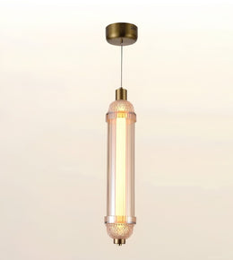 A modern cylindrical pendant light with a warm glowing glass shade and antique brass metal accents, suspended against a soft beige gradient background.