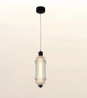 A modern cylindrical pendant light with a ribbed clear glass shade, a warm glowing bulb inside, and black metal hardware, suspended against a soft beige gradient background.