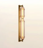 A modern wall-mounted light fixture with two vertical cylindrical glass shades and a brushed gold metal frame, shown against a plain off-white background.