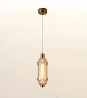 A modern cylindrical pendant light with a textured clear glass shade and brushed brass metal accents, suspended from a matching ceiling canopy against a soft warm gradient background.