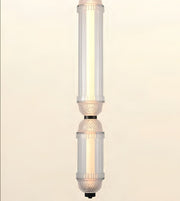 A modern vertical pendant light fixture with two stacked cylindrical glass shades, featuring a warm glowing light inside and textured metallic accents near the middle, set against a solid cream background.