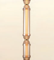 A close-up of a decorative hanging light fixture with clear cylindrical glass shades and warm metallic rose-gold or copper-toned accents, shown against a soft cream background.