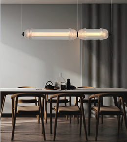 A modern two-light linear pendant fixture with ribbed cylindrical glass shades hangs above a dining table with wooden chairs in a minimalist interior.