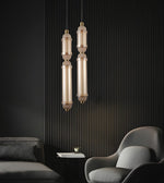 Two modern pendant lights with elongated cylindrical glass shades and brushed brass-toned metal accents, hanging in a dark contemporary room with a ribbed black wall, gray sofa, and curved gray armchair.