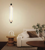 A modern cylindrical pendant light with a glowing vertical tube hangs above a minimalist living room setup featuring a cream sofa, a wooden side table, and a potted plant against a warm beige wall.