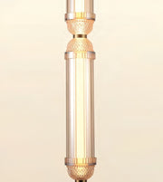 A close-up of a cylindrical pendant light fixture with a warm white glowing bulb inside a clear glass shade, accented by gold-toned metal bands and textured details on a light beige background.