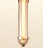 A modern cylindrical pendant light fixture with a warm glowing light inside a clear glass shade, accented by polished gold-toned metal rings and a textured glass finial at the bottom.